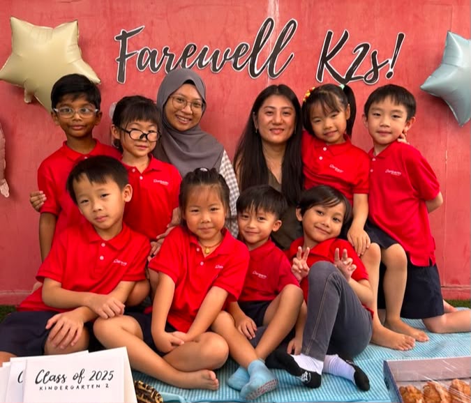 Teachers and Kindergarten 2 graduates posing together in front of a 'Farewell K2s!' banner at their graduation celebration